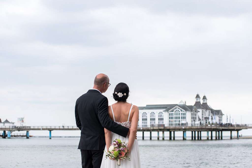 Fotograf Mazelle fotografiert das Brautpaar mit Blick auf die Seebrücke in Sellin an der Ostsee-Hochzeitsfotografie von Mazelle Photography Fotostudio®