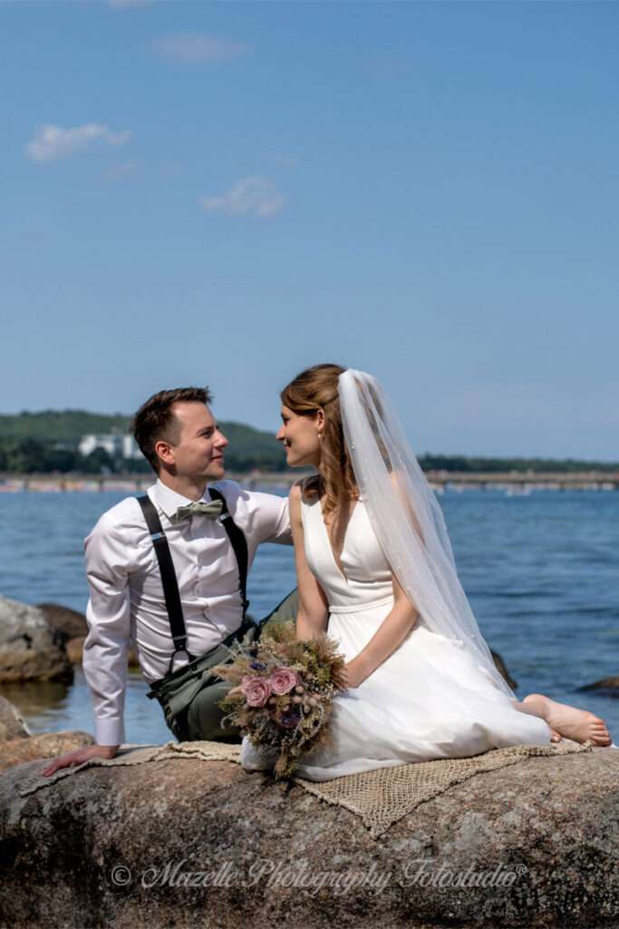 Ein frisch vermähltes Brautpaar sitzt auf einem Stein in der Ostsee vor Binz und schaut sich tief in die Augen – ein Moment voller Ruhe, Nähe und Verbundenheit. Das Hochzeitsfotoshooting fand direkt nach der Trauung im Müther-Turm statt und wurde exklusiv begleitet von den Hochzeitsfotografen des Mazelle Photography Fotostudio®. Die Ostsee, das Licht und die Natürlichkeit des Augenblicks verschmelzen zu einem Bild, das mehr erzählt als Worte.