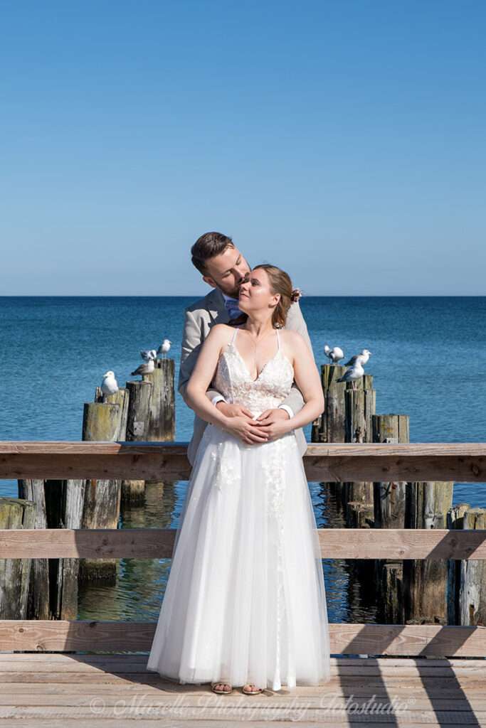 Ein Hochzeitspaar küsst sich innig auf der Seebrücke in Zingst. Die Szene wirkt ruhig und poetisch: Links und rechts sitzen Möwen auf den Holzpfählen, als stille Beobachter des Moments. Die Perspektive ist liebevoll gewählt – eingefangen von den Hochzeitsfotografen Mazelle Photography Fotostudio® von Rügen, die mit viel Gefühl und Präzision arbeiten. Das Bild verbindet Natur, Emotion und Komposition zu einem ausdrucksstarken Hochzeitsmoment an der Ostsee.