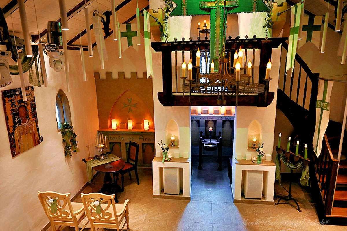 Der Innenraum der Ordenskapelle Binz zeigt eine festliche und intime Atmosphäre: Zwei Stühle für das Hochzeitspaar, ein Tisch in Form eines Ritterschildes mit Kerzenleuchter auf grüner Tischdecke sowie ein Schieferherz, auf dem die Eheringe bis zum Ringtausch liegen. Hochzeitsfotograf Mazelle hält diese besonderen Details kunstvoll fest.