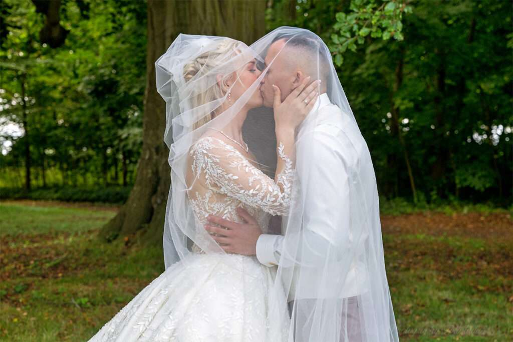 Beim Hochzeitsshooting im Schlosspark des Schlosses Hohendorf küsst sich das Brautpaar innig unter dem Brautschleier, direkt unter einer großen Eiche. Der hochzeitsfotograf des mazelle photography fotostudio® aus dranske auf rügen hat diesen romantischen Moment eingefangen. Die natürliche Kulisse des Schlossparks, kombiniert mit der Symbolik des Brautschleiers, verleiht den Hochzeitsfotos eine besondere Eleganz und Authentizität. So entstehen Bilder, die die Liebe und Verbundenheit des Paares eindrucksvoll sichtbar machen und als wertvolle Erinnerung bleiben.